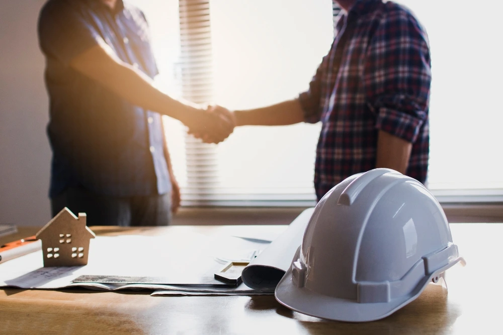 Two-professionals-shaking-hands-in-an-office-setting-with-a-hard-hat-and-plans-on-the-table